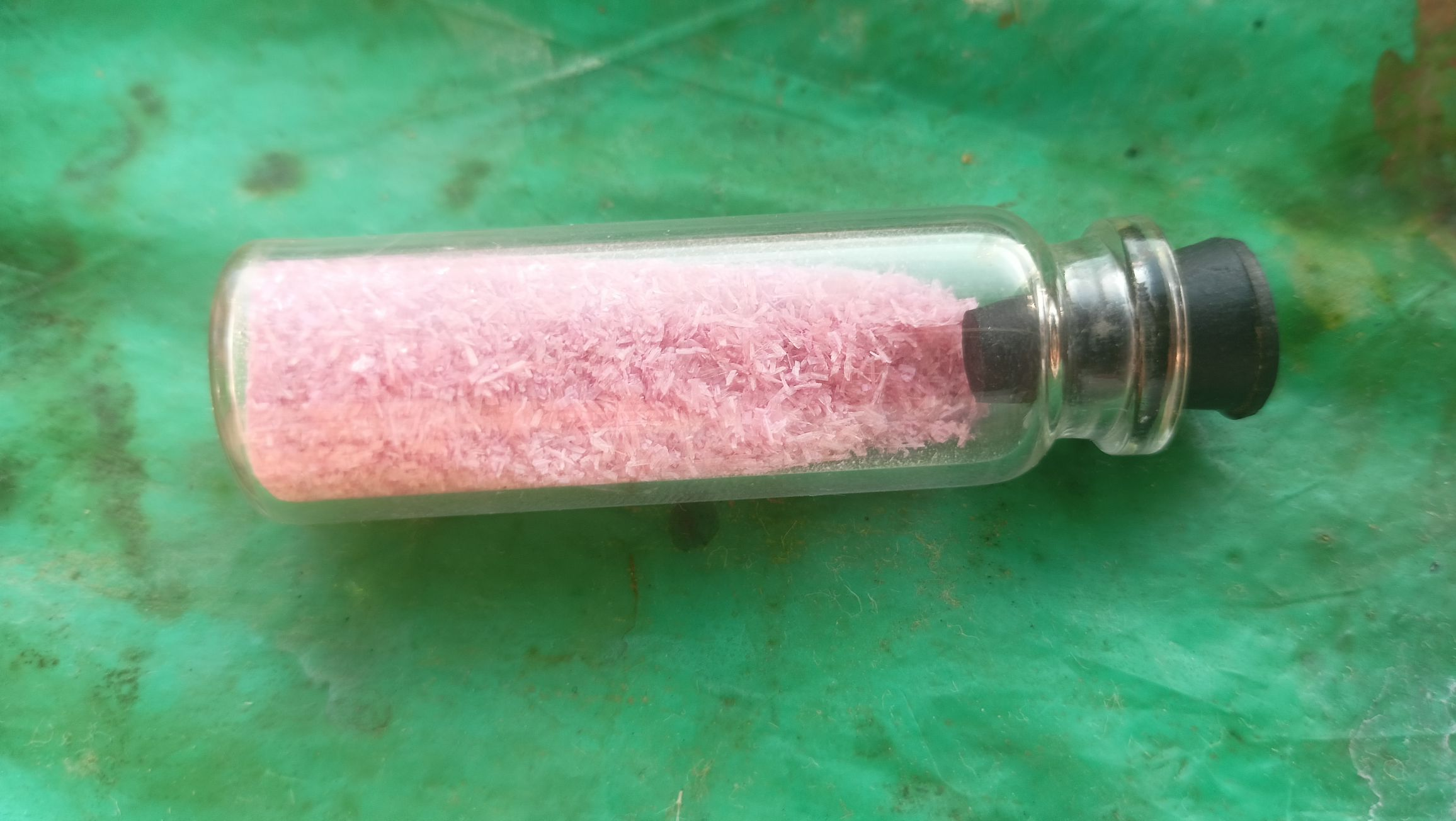 Pink Needle-like crystals in a vial stoppered by a black rubber stopper on a green background